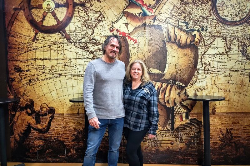 Brewery owners Rick and Cynthia Lee stand in front of an interior wall decorated with pirate ships inside DarkHeart Brewing